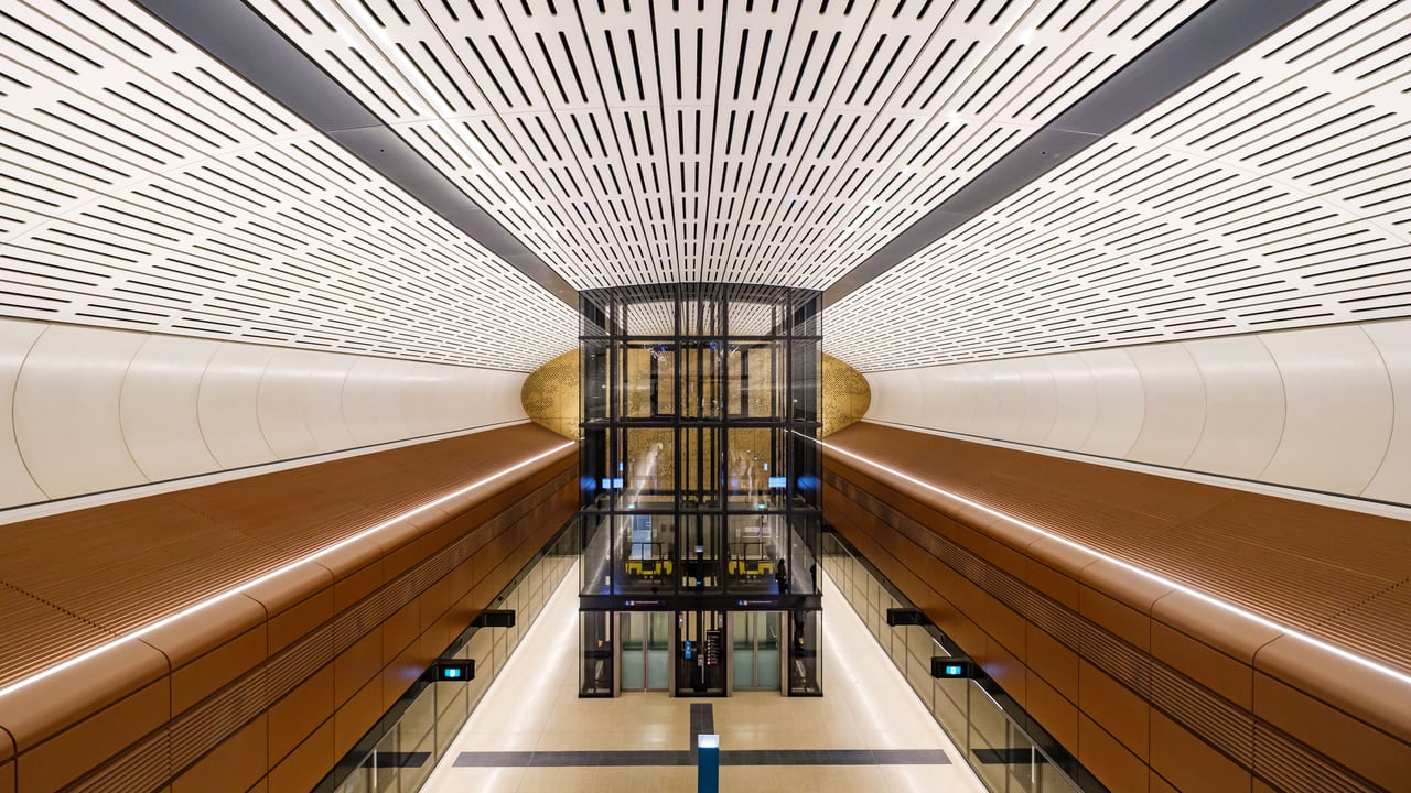 The platform level of a new Sydney Metro station, a glass lift shaft rising between timber-panelled walls beneath a vaulted ceiling