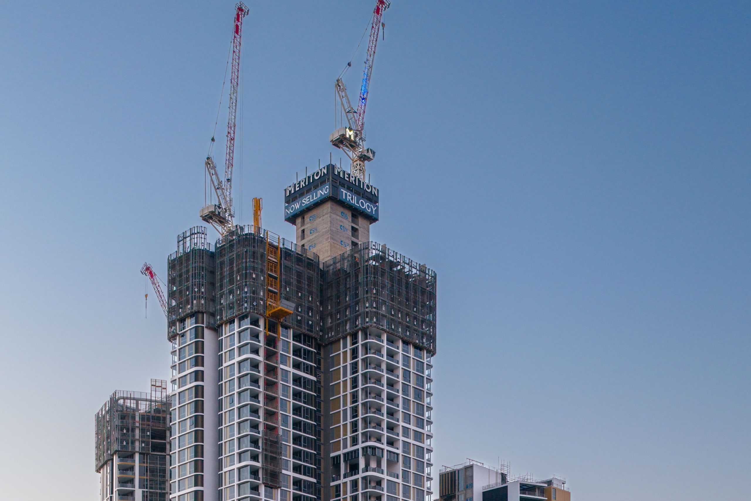 A residential apartment tower under construction against a clear Sydney sky, tower cranes rising over the part-built concrete frame